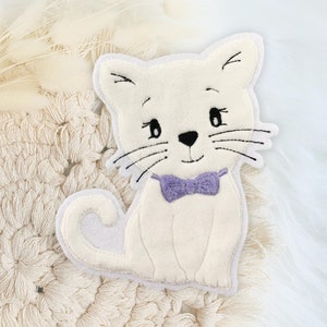 Könnte beinhalten: Ein weißer Plüsch-Katzen-Applik mit gestickten Details, darunter eine violette Schleife. Der Applik liegt auf einer cremefarbenen gehäkelten Oberfläche.
