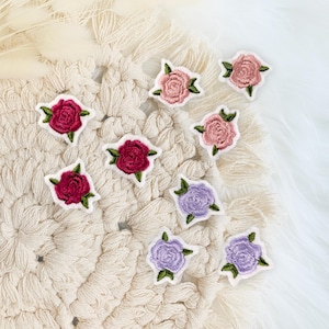 Könnte beinhalten: Acht gestickte Rosen-Patches in verschiedenen Farben, darunter Rosa, Lila und Rot, sind auf einer cremefarbenen gehäkelten Oberfläche angeordnet.