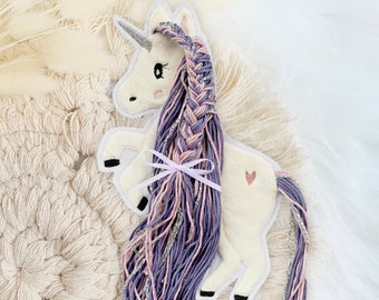 Puede incluir: Un unicornio de fieltro blanco con una crin y una cola trenzadas de color morado y rosa. El unicornio tiene un cuerno plateado y un corazón rosa en la espalda.