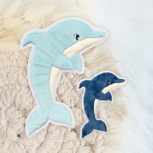 Könnte beinhalten: Zwei Plüsch-Delphin-Spielzeuge. Eines ist hellblau mit weißem Bauch und grauem Umriss, das andere ist dunkelblau mit weißem Bauch. Beide haben schwarze Augen und liegen auf einer strukturierten, cremefarbenen Oberfläche.