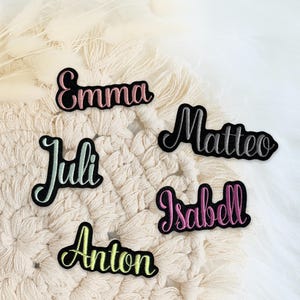Könnte beinhalten: Fünf einzelne bestickte Namensaufnäher mit schwarzem Rand. Die Namen Emma, Matteo, Juli, Isabell und Anton sind in Schreibschrift mit verschiedenen Garnfarben gestickt, darunter Rosa, Silber, Hellgrün, Violett und Gelb.