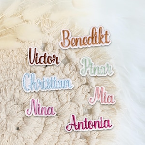 Könnte beinhalten: Eine Sammlung von sieben bestickten Namensaufnähern mit den Namen Benedikt, Victor, Pinar, Christian, Mia, Nina und Antonia. Jeder Aufnäher ist in Schreibschrift gestickt und weist verschiedene Garnfarben wie Braun, Hellgrün, Blau, Rosa und Magenta auf.
