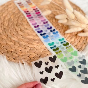 Könnte beinhalten: Herzförmige Aufkleber in verschiedenen Farben, darunter Schwarz, Rosa, Blau und Grün. Die Aufkleber sind auf transparenten Bögen angeordnet, einige werden von einer Hand gehalten. Der Hintergrund umfasst eine gewebte Matte und flauschigen weißen Stoff.