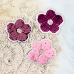 Könnte beinhalten: Drei Filzblumen-Patches in Pink, Burgunderrot und Hellrosa mit goldenen Glitzerakzenten. Die Patches befinden sich auf einer cremefarbenen gehäkelten Oberfläche.