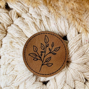 Könnte beinhalten: Ein brauner Lederuntersetzer mit einem genähten Rand und einer schwarzen Umrandung eines Blatt-Zweig-Designs.