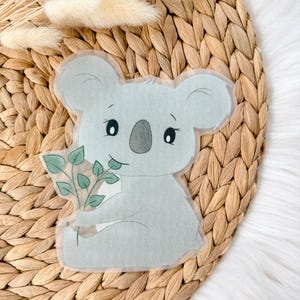 Könnte beinhalten: Ein hellblauer Koala, der einen Zweig mit grünen Eukalyptusblättern hält. Der Koala sitzt auf einem geflochtenen braunen Hintergrund.