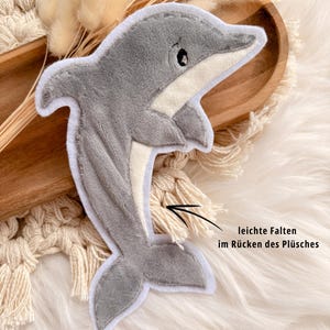 Könnte beinhalten: Ein grauer Plüschdelfin mit weißem Bauch und Besatz. Der Delfin hat ein kleines schwarzes Auge und liegt auf einer weißen, flauschigen Oberfläche. Es ist ein Stofftier.