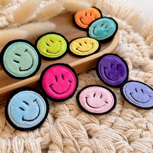 Könnte beinhalten: Eine Sammlung bunter Smiley-Aufnäher. Jeder Aufnäher hat eine andere Farbe, darunter Rosa, Blau, Grün, Gelb, Orange und Lila, mit einem schwarzen Rand und einem lächelnden Gesichtsdesign. Die Aufnäher sind rund und aus einem weichen, strukturierten Material.