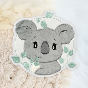 Könnte beinhalten: Ein grauer Koalabär mit schwarzen Augen und einem kleinen Mund ist auf einen weißen Stoffkreis gestickt. Der Koala ist von grünen Blättern umgeben.