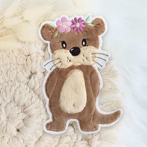 Könnte beinhalten: Gestickter Aufnäher mit einem Cartoon-Otter mit Blumenkranz. Der Otter ist braun mit cremefarbenem Bauch und weißer Umrandung. Die Blumen sind rosa und lila. Der Aufnäher ist für das Aufbringen auf Kleidung oder Accessoires gedacht.