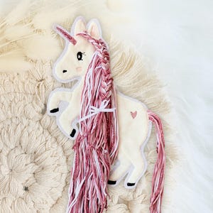 Könnte beinhalten: Ein weißer Filz-Einhorn-Applikator mit rosa und weißem Garnhaar, einem rosa Herz an der Seite und einem geflochtenen rosa und weißen Garn-Schwanz.