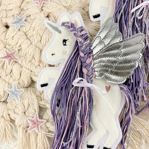 May include: A white felt unicorn with silver wings and a purple and pink braided mane. The unicorn is on a white crocheted background with pink and silver stars.