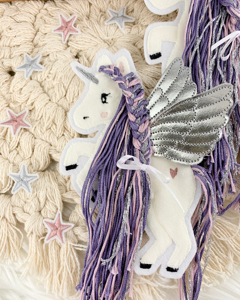 May include: A white felt unicorn with silver wings and a purple and pink braided mane. The unicorn is on a white crocheted background with pink and silver stars.