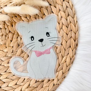 Könnte beinhalten: Eine graue Katze mit einer rosa Schleife sitzt auf einer gewebten Oberfläche. Die Katze hat einen weißen Schwanz und schwarze Schnurrhaare.