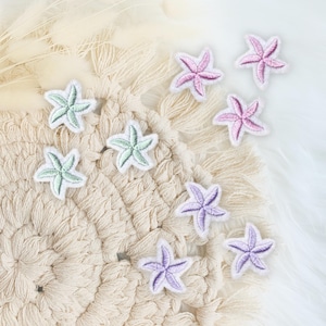 Könnte beinhalten: Eine Ansammlung von gestickten Seestern-Patches in Rosa, Lila und Grün mit weißen Rändern. Die Patches liegen auf einer strukturierten beigefarbenen Oberfläche und einem flauschigen weißen Hintergrund.