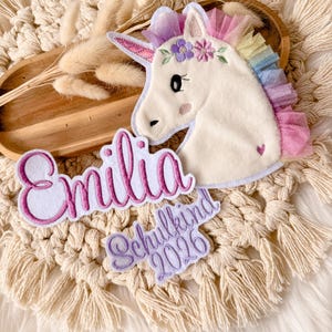 Op de afbeelding: Een witte eenhoorn applicatie met een regenboog manen en een bloemenkrans. De naam "Emilia" is geborduurd in roze script, samen met "Schulkind 2026" in paars. De applicatie is geplaatst op een macrame achtergrond.