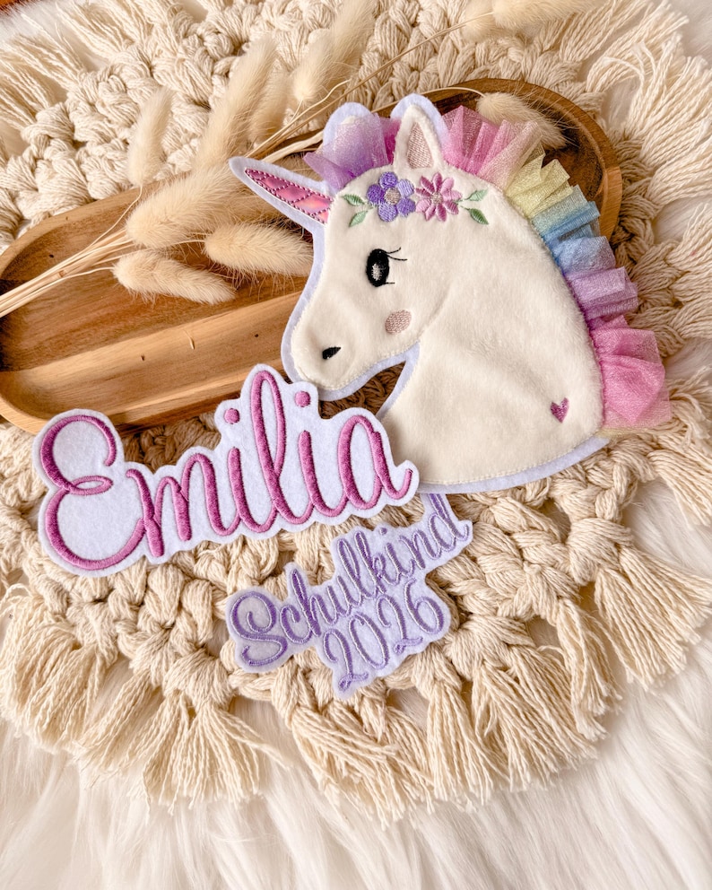 Op de afbeelding: Een witte eenhoorn applicatie met een regenboog manen en een bloemenkrans. De naam "Emilia" is geborduurd in roze script, samen met "Schulkind 2026" in paars. De applicatie is geplaatst op een macrame achtergrond.