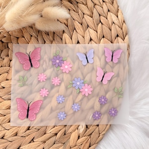 Könnte beinhalten: Eine transparente, rechteckige Folie mit rosa und lila Schmetterlingen und Blumen. Die Schmetterlinge haben schwarze Akzente. Die Blumen sind in Rosa-, Lila- und Blautönen gehalten, mit grünen Blättern. Auf einer hellbraunen, gewebten Oberfläche.