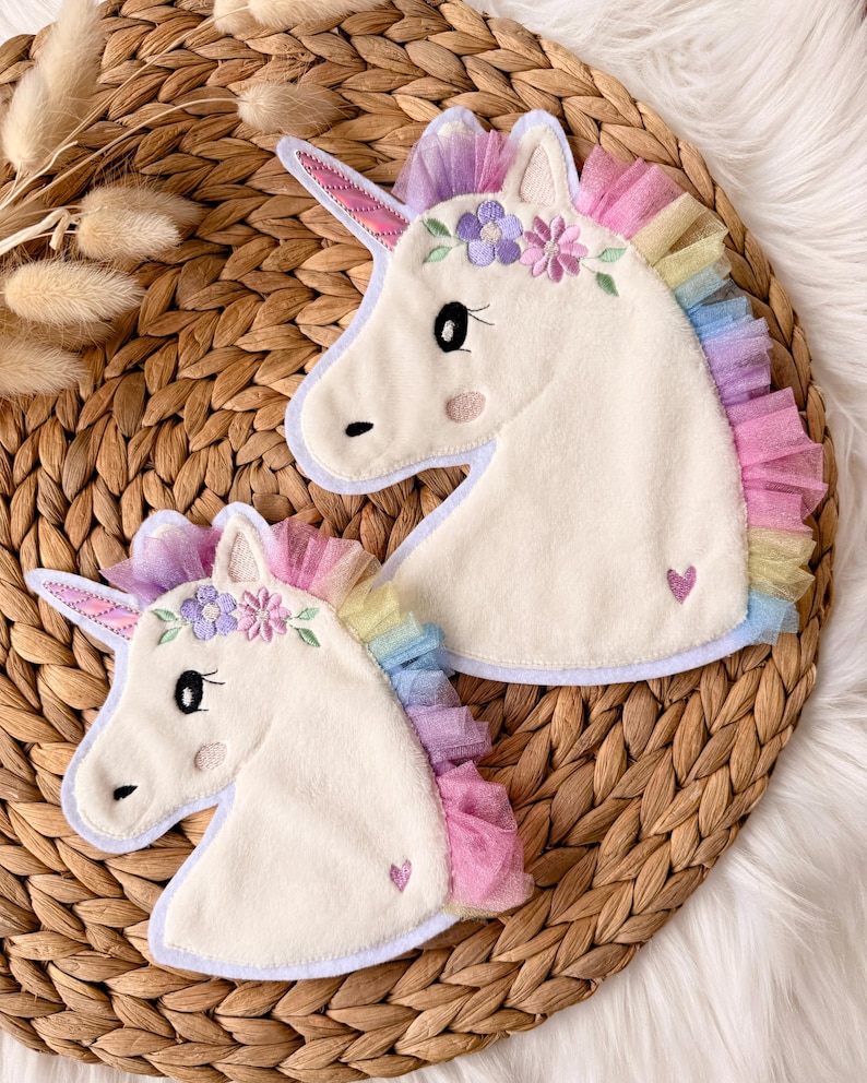 Op de afbeelding: Twee witte eenhoornvormige applicaties met kleurrijke manen en bloemaccenten. De applicaties hebben een hartdetail en zijn geplaatst tegen een geweven achtergrond. De manen hebben tinten roze, geel, blauw en paars.