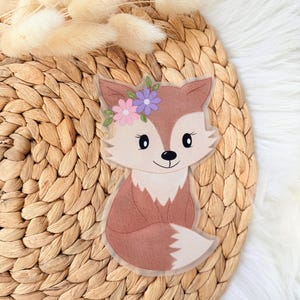 Könnte beinhalten: Ein skurriler Fuchs-Aufkleber mit einem Blumenkranz. Der Aufkleber zeigt einen braunen Fuchs mit weißer Brust und Schwanz sowie einen Blumenkranz mit rosa und lila Blumen. Der Aufkleber befindet sich auf einer gewebten, beigefarbenen Oberfläche.