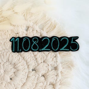 Könnte beinhalten: Ein schwarzer und türkisfarbener gestickter Aufnäher mit dem Datum "11.08.2025" auf einem weißen gehäkelten Hintergrund.