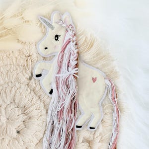 Könnte beinhalten: Ein weißes Filz-Einhorn mit einer pinkfarbenen und weißen geflochtenen Mähne und einem Schwanz. Das Einhorn hat ein kleines pinkfarbenes Herz auf der Seite gestickt.