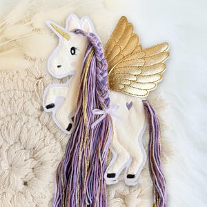 Könnte beinhalten: Ein weißes Filz-Einhorn mit goldenem Horn und Flügeln. Das Einhorn hat eine lange, geflochtene Mähne und einen Schweif in Lila- und Goldtönen. Ein kleines lila Herz befindet sich auf dem Körper des Einhorns. Ein skurriles Deko-Objekt.