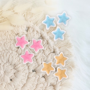 Könnte beinhalten: Nahaufnahme einer beigefarbigen Makramee-Wandbehang mit sechs Filzsternen in verschiedenen Farben: rosa, hellblau und gelb. Die Sterne sind in einer Gruppe auf dem Makramee angeordnet.