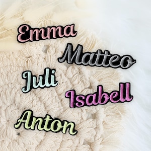 Könnte beinhalten: Fünf bestickte Namensaufnäher mit schwarzem Rand und kursiver Schrift. Die Namen sind Emma in Pink, Matteo in Silber, Juli in Hellblau, Isabell in Magenta und Anton in Hellgrün, alle mit einer deutlich sichtbaren gestickten Textur.