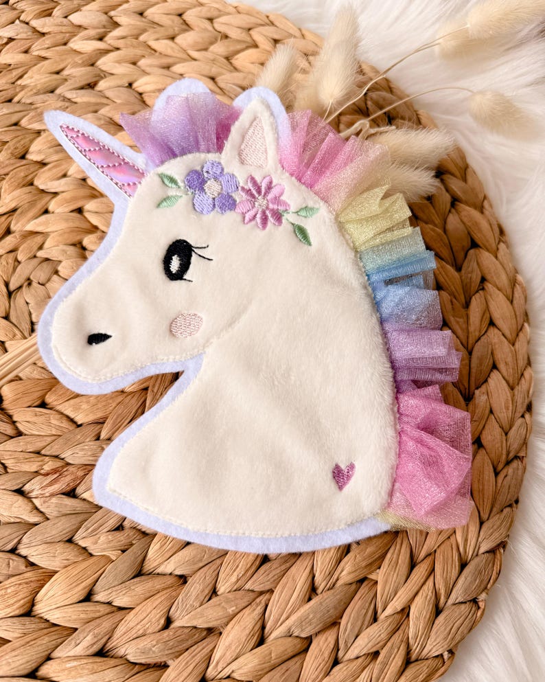 Op de afbeelding: Een pluche eenhoofd met een witte body, versierd met een paarse en roze bloemenkrans en een regenboogmanen. De eenhoorn heeft een klein roze hartje en een lavendelkleurige rand.