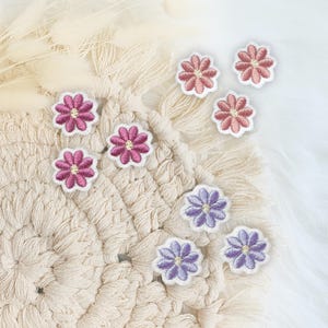 Könnte beinhalten: Sortiment an gestickten Blumen-Patches in den Farben Pink, Lila und Lavendel. Die Patches sind auf einer weißen gehäkelten Oberfläche angeordnet.