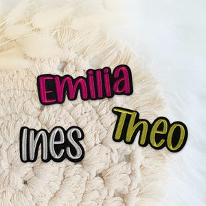 Könnte beinhalten: Drei bestickte Namensaufnäher mit schwarzem Rand. Der obere Aufnäher zeigt den Namen Emilia in leuchtendem Pink, der untere linke Ines in Silber und der untere rechte Theo in Metallic-Gold. Jeder Name ist in einer Schreibschrift gehalten.