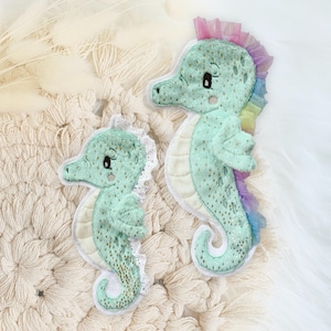 Könnte beinhalten: Zwei gestickte Seepferdchen-Aufnäher in blaugrün mit goldenen Glitzerakzenten. Das größere Seepferdchen hat einen Regenbogen-Tüllbesatz.