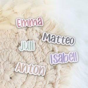 Könnte beinhalten: Fünf einzelne Stoffaufnäher mit gestickten Namen in verschiedenen Farben. Die Aufnäher zeigen die Namen Emma in Rosa, Matteo in Grau, Juli in Hellgrün, Isabell in Helllila und Anton in Beige, jeweils mit einem weißen Rand versehen.