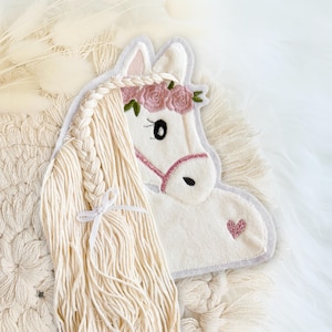Könnte beinhalten: Weißer Filz-Einhornkopf mit rosa Blumenkranz und rosa Herz. Das Einhorn hat eine lange, geflochtene Mähne und einen rosa Zaum. Der Hintergrund ist eine strukturierte, cremefarbene Oberfläche.