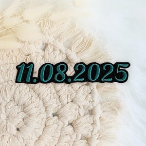 Könnte beinhalten: Ein gestickter Patch in Tealblau und Schwarz mit dem Datum "11.08.2025" auf einem weißen Häkelhintergrund.