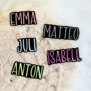 Könnte beinhalten: Fünf rechteckige schwarze Stoffaufnäher mit gestickten Namen in verschiedenen Farben. Die Namen EMMA, MATTEO, JULI, ISABELL und ANTON sind in rosa, grau, hellblau, lila und lindgrünem Garn aufgestickt.