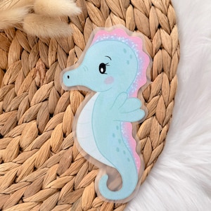 Könnte beinhalten: Ein hellblauer Seepferdchen-Aufkleber mit rosa und lila Flossendetails. Der Aufkleber hat ein Cartoon-Design mit einem lächelnden Gesicht und ist auf einer gewebten Korb-Oberfläche platziert.