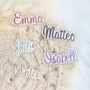 Könnte beinhalten: Fünf gestickte Patches mit verschiedenen Namen: Emma, Matteo, Juli, Isabell und Anton. Die Patches befinden sich auf einer weißen gehäkelten Oberfläche.
