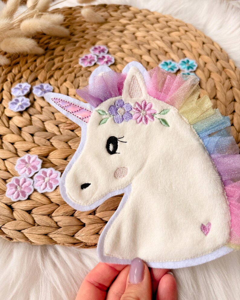 Op de afbeelding: Een witte eenhoorn applicatie met een regenboog manen en een bloemenkrans. De eenhoorn heeft een roze hart en een blush-kleurige wang. Verschillende bloemvormige patches in roze en paarse tinten zijn rond de eenhoorn verspreid.