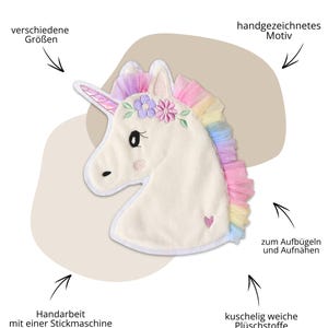 Op de afbeelding: Een cr&egrave;me-kleurige pluche eenhoofd met een regenboogmanen en een roze hoorn. De eenhoorn heeft geborduurde details, waaronder een bloemenkrans en een klein roze hart. De afbeelding bevat Duitse tekst.