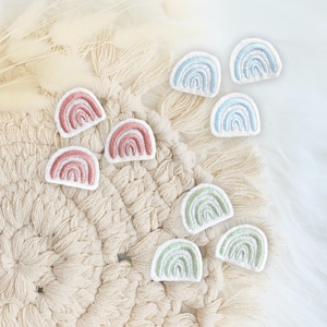 Könnte beinhalten: Gestickte Regenbogen-Patches in Pastellfarben wie Rosa, Blau und Grün. Die Patches sind auf einer cremefarbenen Makramee-Oberfläche und einem weißen, flauschigen Hintergrund angeordnet.