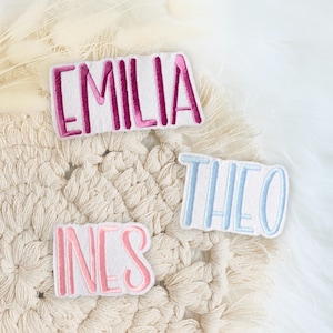 Könnte beinhalten: Drei gestickte Stoff-Patches mit den Namen "Emilia", "Theo" und "Ines" in rosa, blau und rosa Schrift. Die Patches befinden sich auf einer weißen, flauschigen Oberfläche.