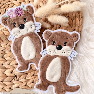 Könnte beinhalten: Zwei braune Otter-förmige Aufnäher mit weißen Akzenten. Ein Otter hat einen Blumenkranz. Beide haben schwarze Augen, Nasen und weiße Schnurrhaare. Die Aufnäher befinden sich auf einem gewebten Hintergrund mit getrockneten Blumen und einer weißen, flauschigen Oberfläche.