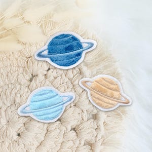 Könnte beinhalten: Drei gestickte Filzpatches von Planeten mit Ringen. Die Planeten sind blau, hellblau und beige. Die Ringe sind weiß.