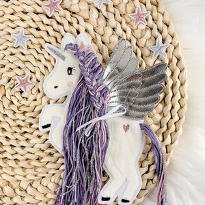 May include: A white felt unicorn with purple and pink yarn hair and silver wings. The unicorn is sitting on a woven straw surface with pink and silver stars.