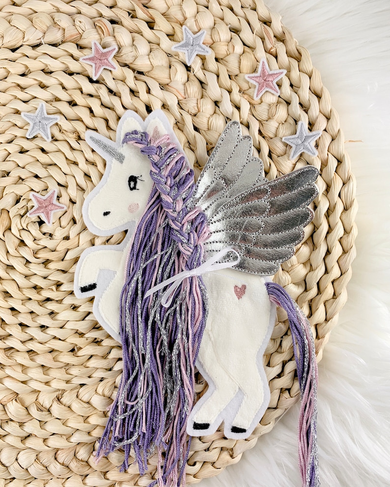 May include: A white felt unicorn with purple and pink yarn hair and silver wings. The unicorn is sitting on a woven straw surface with pink and silver stars.