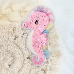 Könnte beinhalten: Ein rosa Seepferdchen-Plüschtier mit einer regenbogenfarbenen Mähne und Schwanz. Das Seepferdchen hat schwarze Punkte und einen weißen Bauch. Das Spielzeug liegt auf einem cremefarbenen Makramee-Hintergrund.