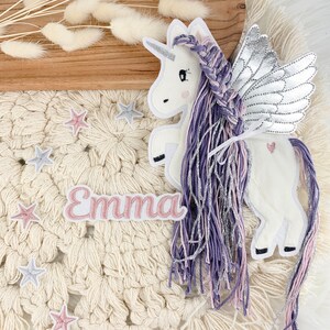 May include: A white unicorn with purple and pink braided hair and silver wings. The unicorn is on a cream-colored crocheted background with pink and silver stars and the name "Emma" in pink letters.