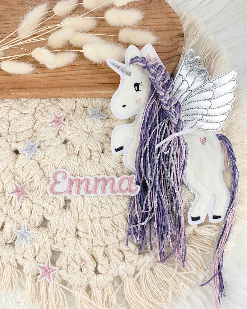May include: A white unicorn with purple and pink braided hair and silver wings. The unicorn is on a cream-colored crocheted background with pink and silver stars and the name "Emma" in pink letters.